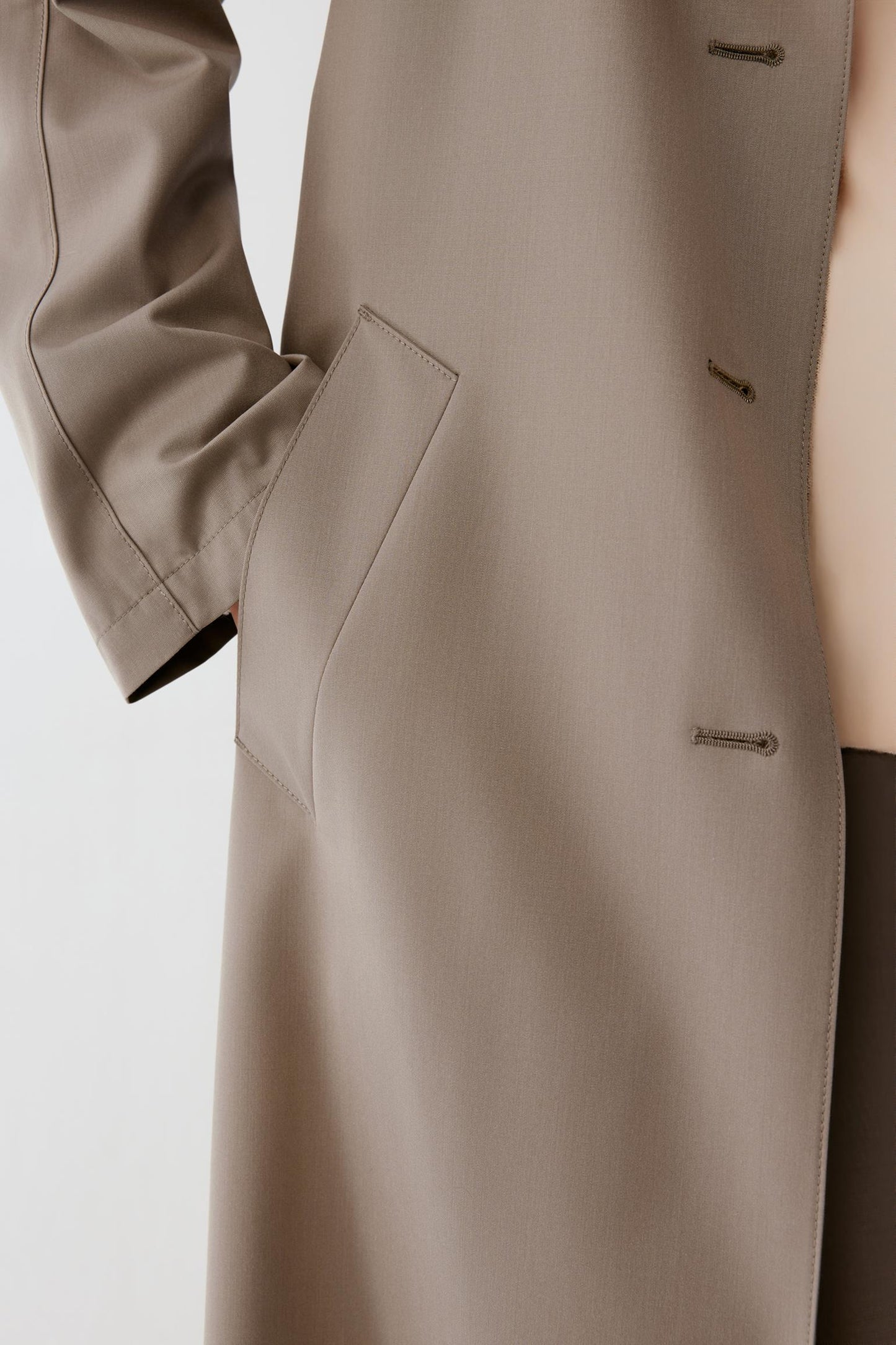 Close-up of a taupe-beige coat with a single-breasted button closure and a side pocket, shown on a model against a light gray background.