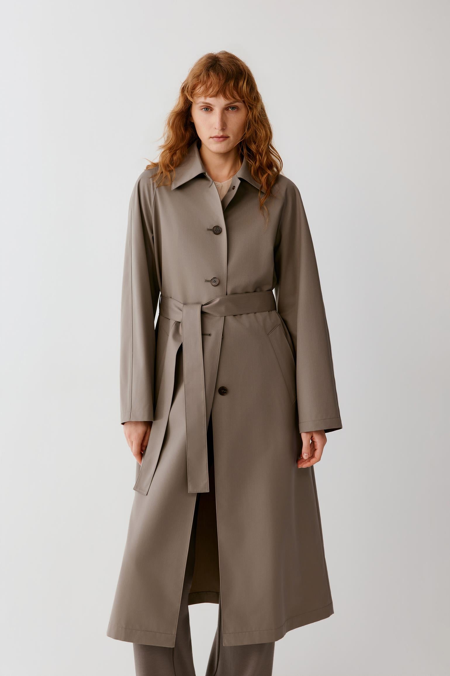 A woman modeling a long taupe-brown trench coat with a matching waist belt, photographed against a plain light gray studio background.