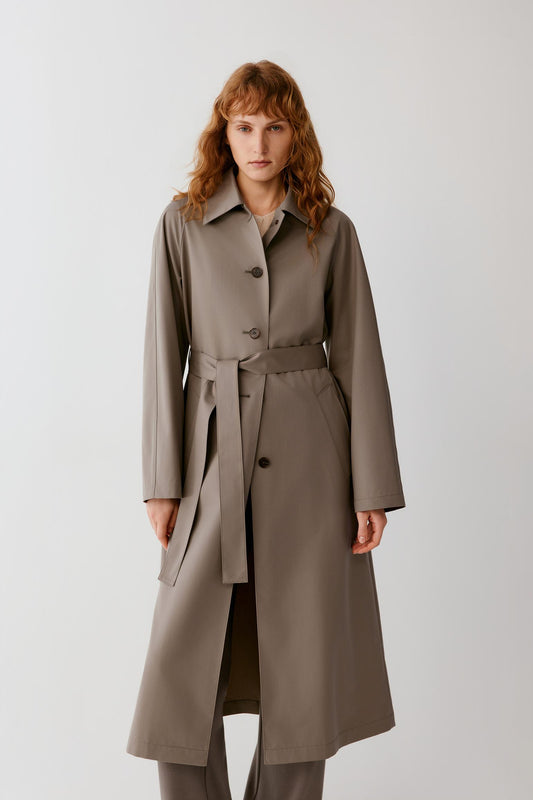 A woman modeling a long taupe-brown trench coat with a matching waist belt, photographed against a plain light gray studio background.