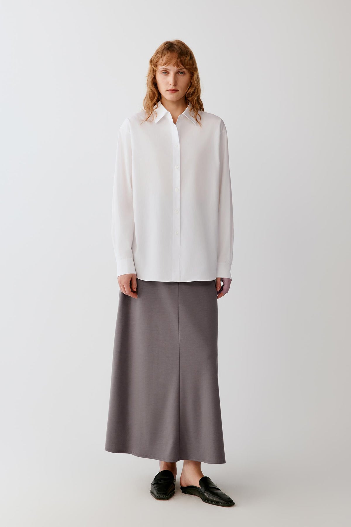 A woman stands against a light gray studio background wearing a loose white long-sleeve button-up shirt and a long taupe-gray skirt, with black loafers.
