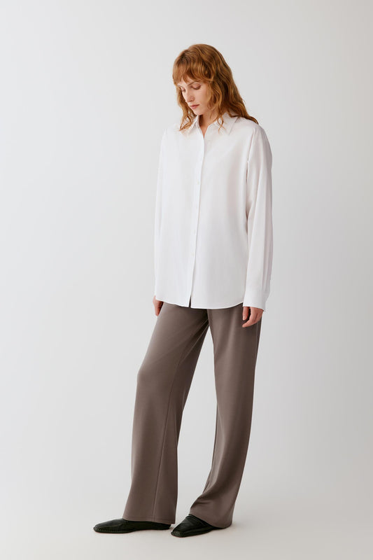 Woman wearing a flowing white shirt and taupe wide-leg trousers against a light grey background.