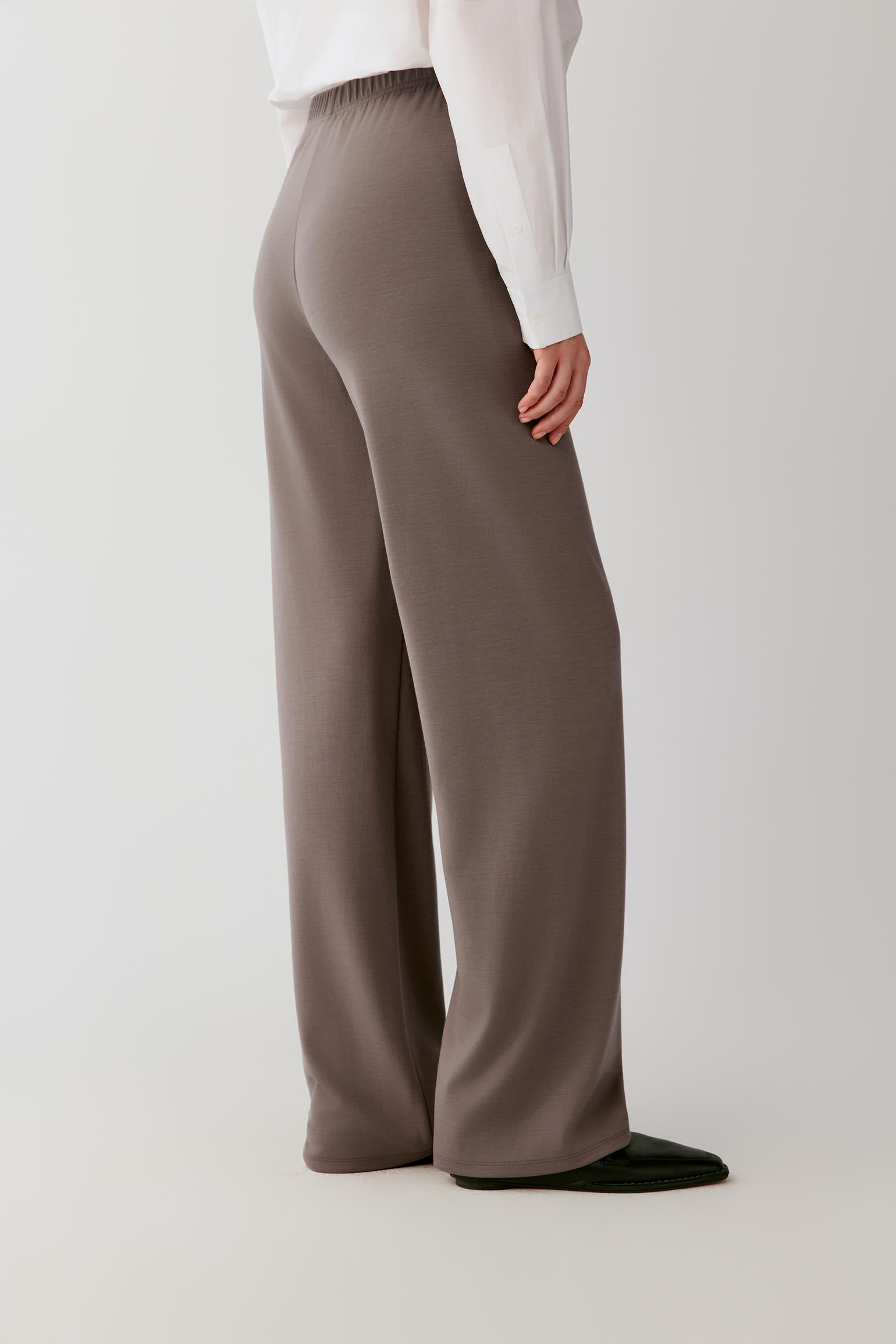 Woman wearing a flowing white shirt and taupe wide-leg trousers against a light grey background.