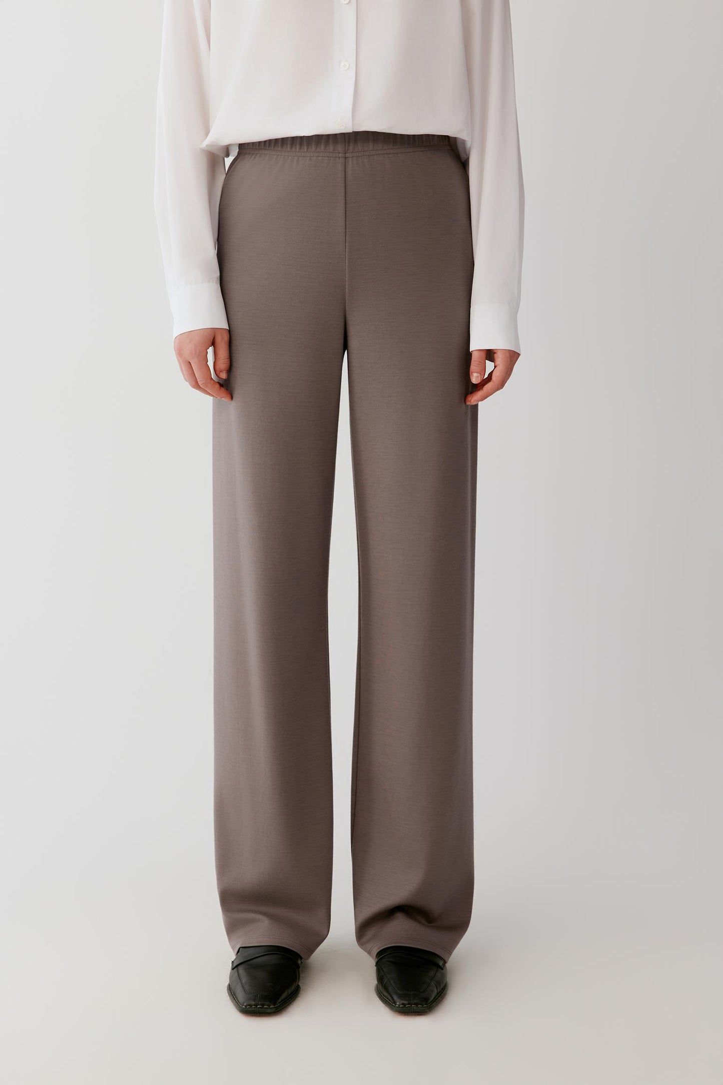 Woman wearing a flowing white shirt and taupe wide-leg trousers against a light grey background.