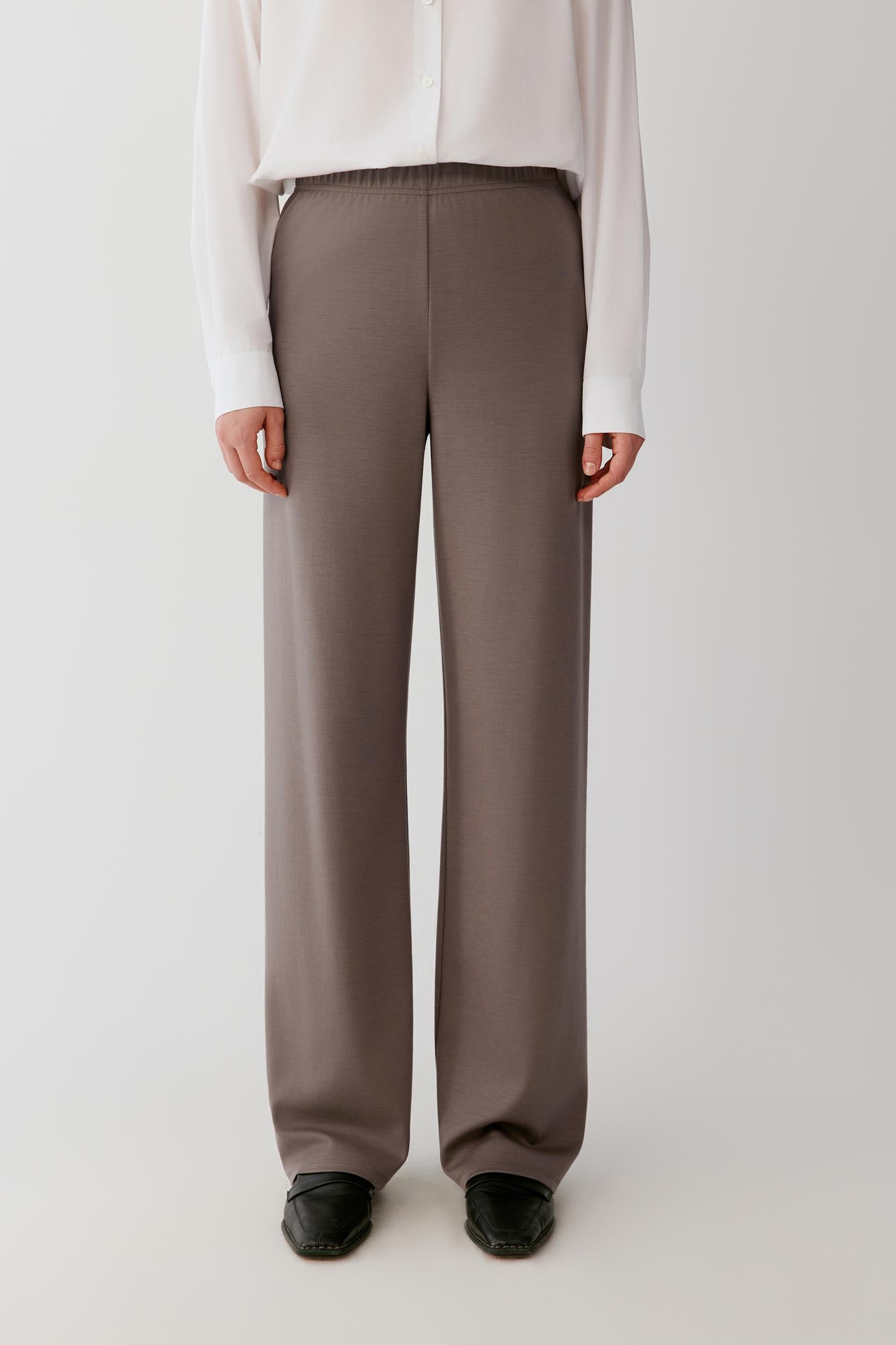 Woman wearing a flowing white shirt and taupe wide-leg trousers against a light grey background.