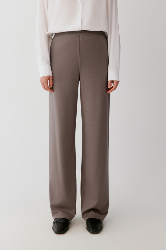 Woman wearing a flowing white shirt and taupe wide-leg trousers against a light grey background.