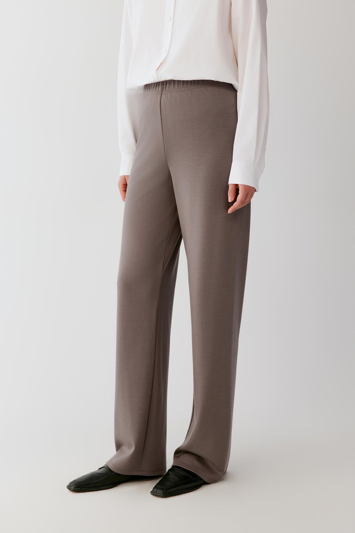 Woman wearing a flowing white shirt and taupe wide-leg trousers against a light grey background.