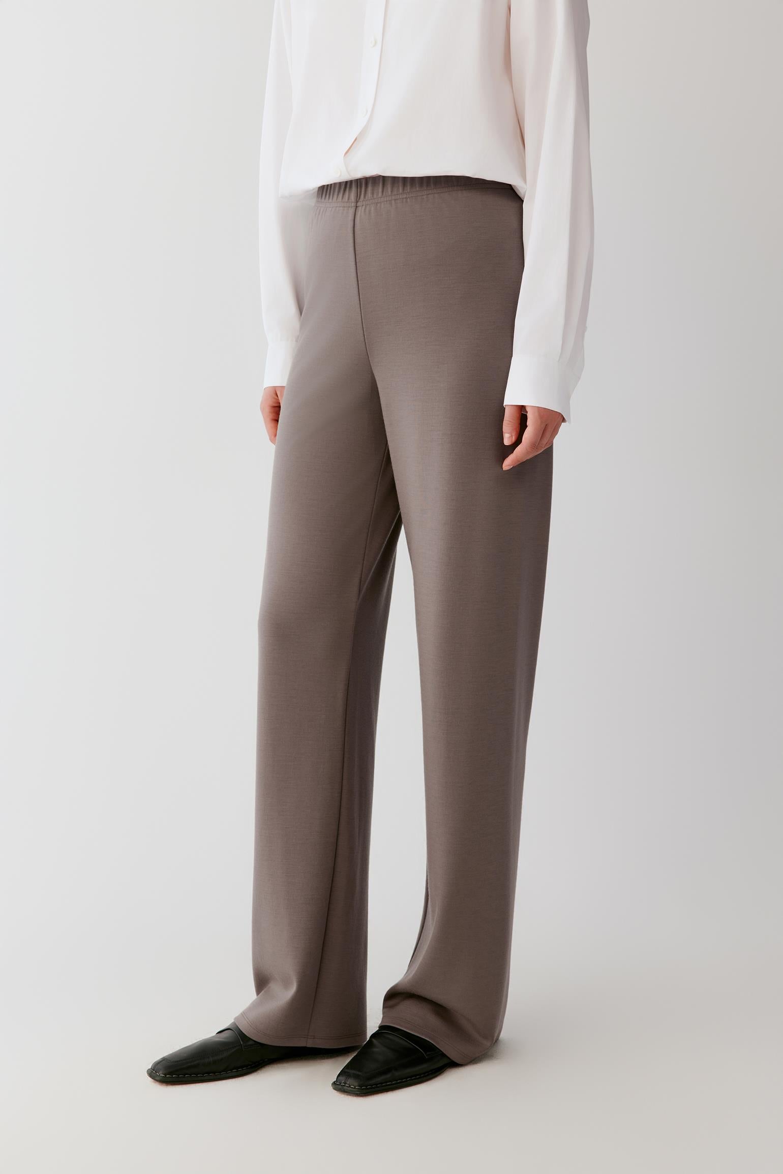 Woman wearing a flowing white shirt and taupe wide-leg trousers against a light grey background.