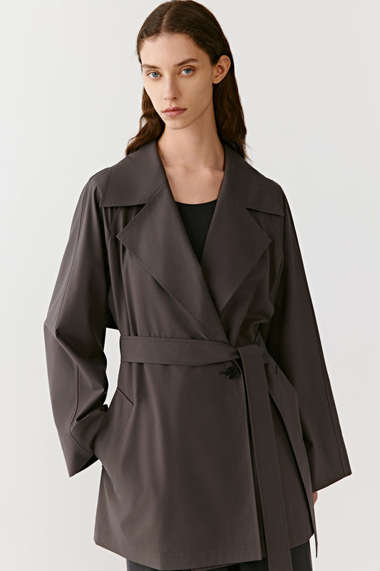 Three-quarter view showing the relaxed straight cut, wide lapels and the belt tied at the waist. The angled side pockets and smooth drape of the fabric are visible.