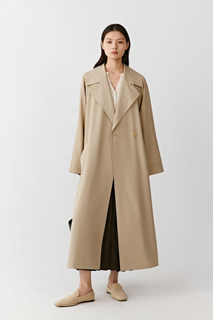Full-length studio photograph of a woman wearing a long beige trench coat in wool with a straight elongated silhouette. The coat falls below the knee and features wide lapels, a concealed front fastening and a matching belt tied loosely at the waist.