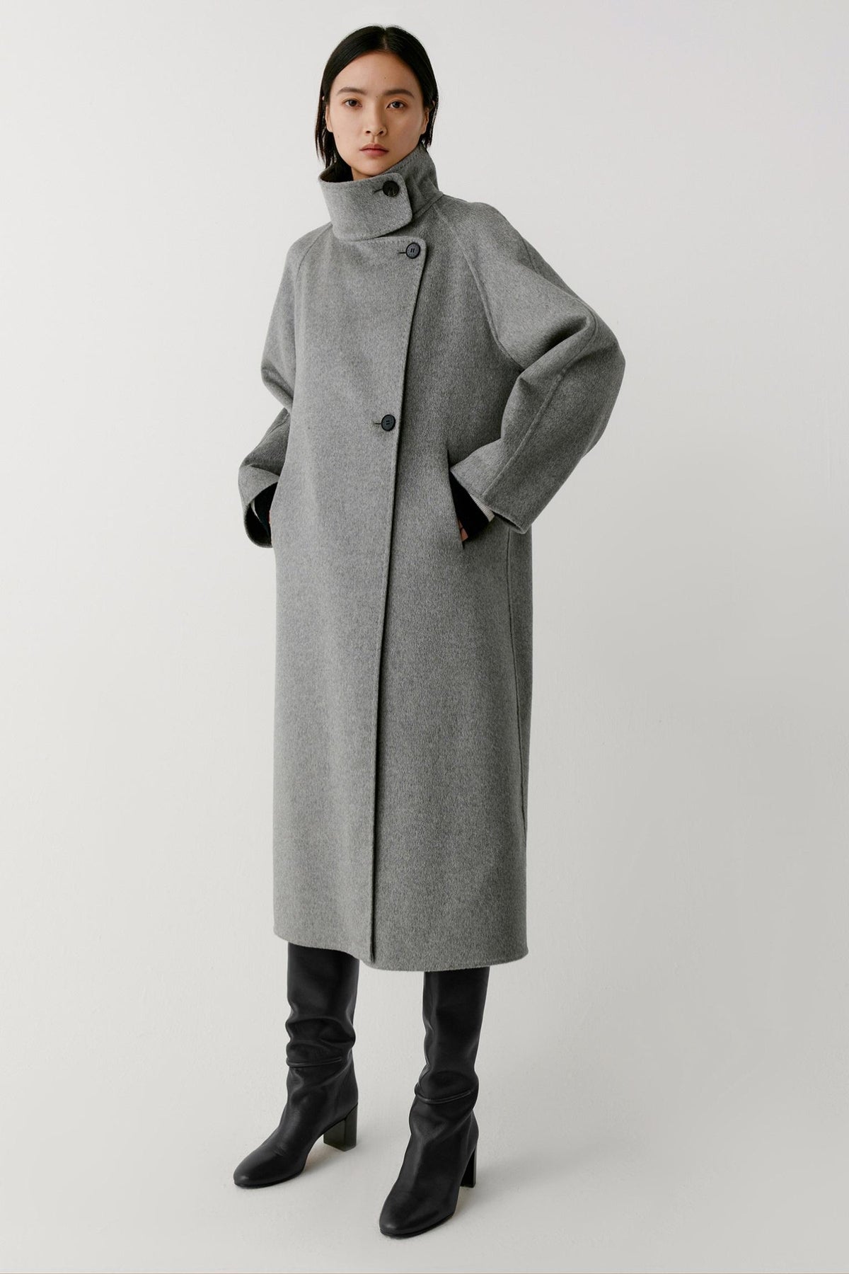 Front view of a woman wearing a grey melange double-face cashmere coat, showing the long silhouette, high stand collar and asymmetric button fastening.