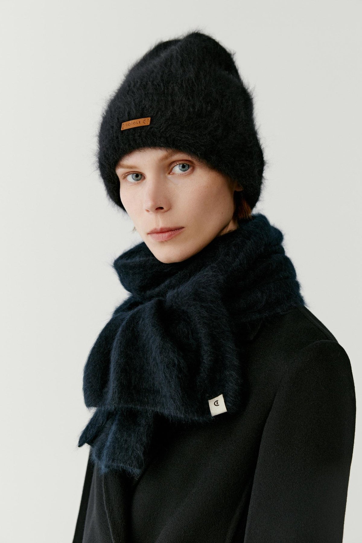 Portrait view of a woman wearing a black cashmere beanie styled with a matching scarf and coat. The image highlights the plush texture, soft volume and fluffy surface of the cashmere knit, while the deep black shade emphasizes the richness and depth of the material. The folded brim and contrasting front label complete the design.