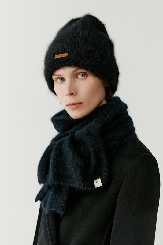 Portrait view of a woman wearing a black cashmere beanie styled with a matching scarf and coat. The image highlights the plush texture, soft volume and fluffy surface of the cashmere knit, while the deep black shade emphasizes the richness and depth of the material. The folded brim and contrasting front label complete the design.