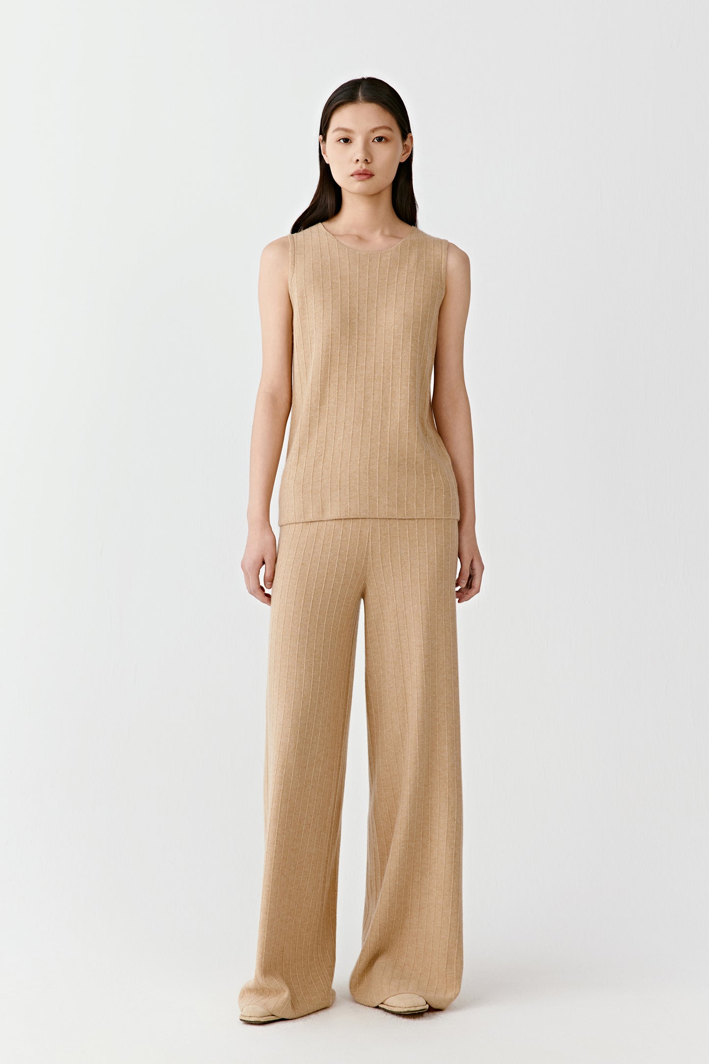 Woman wearing a camel fine ribbed cashmere tank top with a fitted, sleeveless cut, paired with matching trousers; full-length front view highlighting the vertical ribbed texture, clean neckline and close fit through the body.