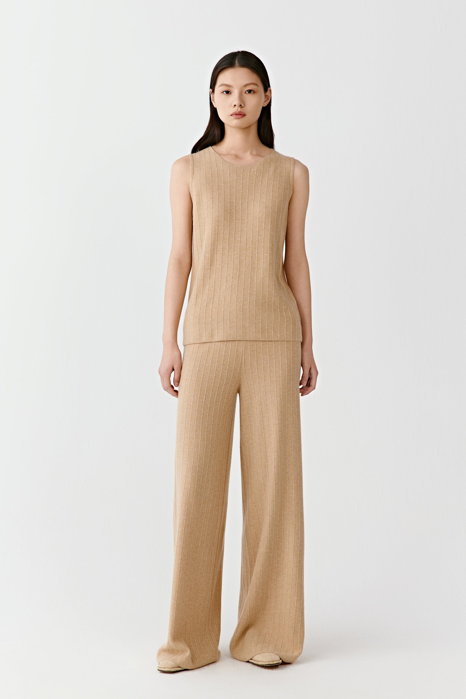 Woman wearing a camel fine ribbed cashmere tank top with a fitted, sleeveless cut, paired with matching trousers; full-length front view highlighting the vertical ribbed texture, clean neckline and close fit through the body.