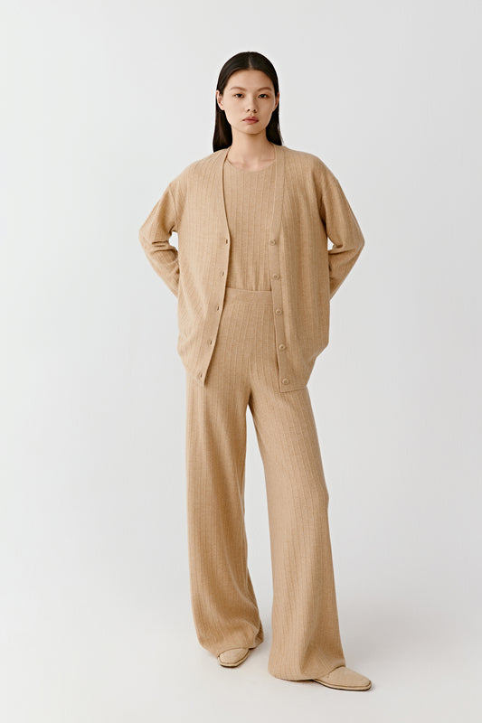 Woman wearing a camel fine rib-knit cashmere cardigan with a straight, relaxed fit and V-neckline, paired with matching trousers; full-length front view highlighting the vertical ribbed texture, button fastening and fluid drape through the body.