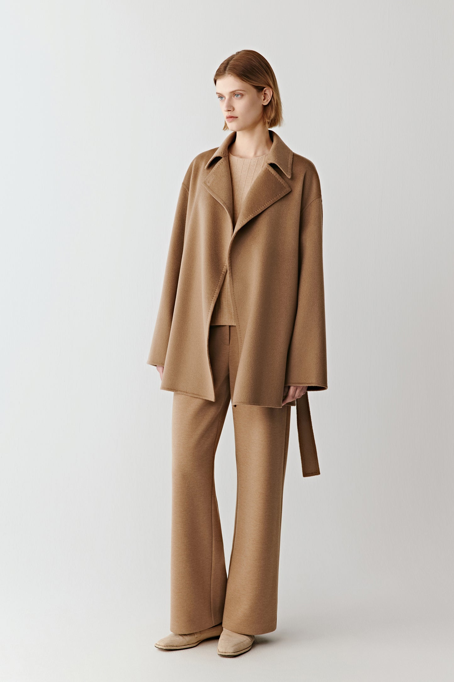 A woman stands facing forward wearing light brown melange straight-leg trousers in cotton, silk and combed wool jersey, paired with a matching coat.