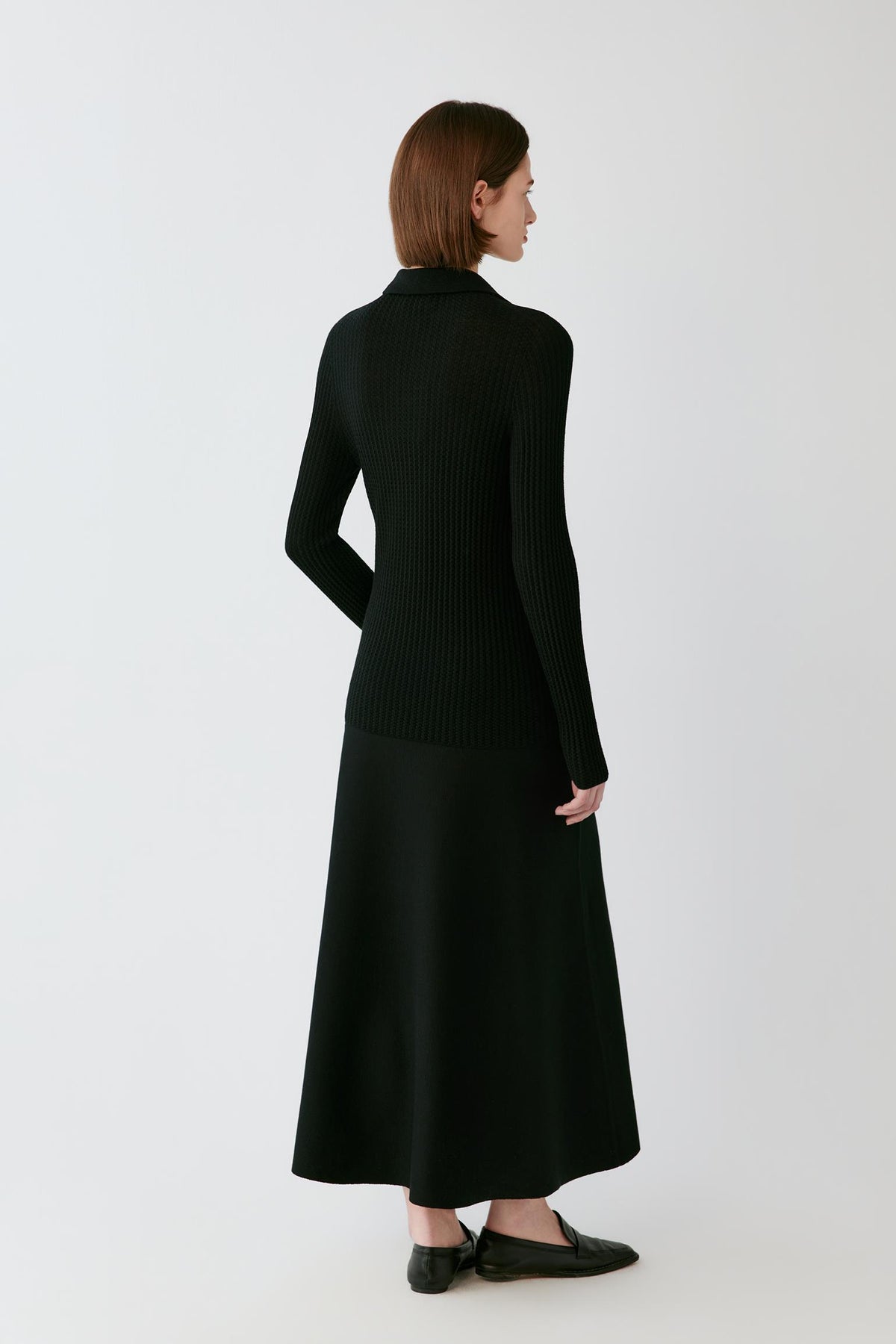 Back full-length view of a woman wearing the deep black worsted wool pullover, showing the straight back and ribbed knit construction.