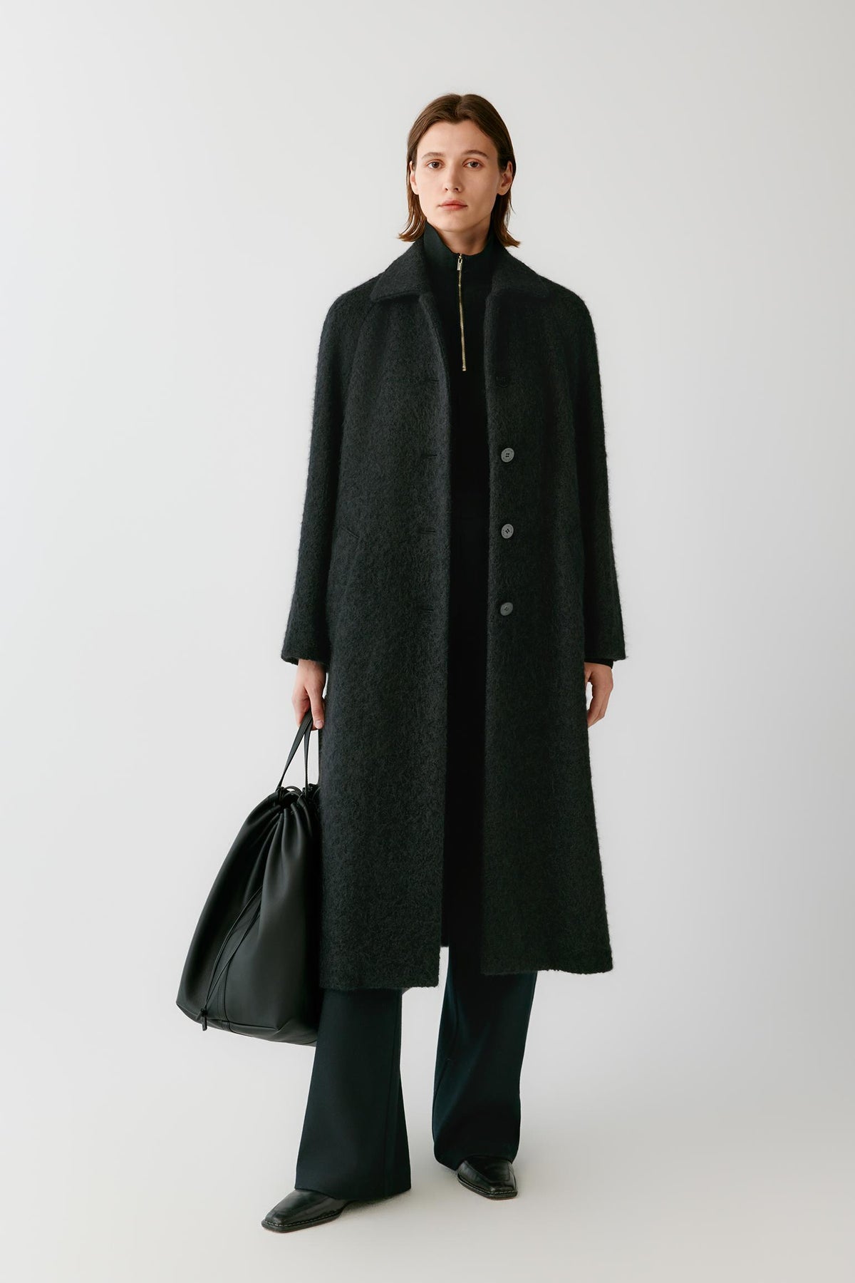 Front view of a woman wearing a deep black alpaca and wool tweed coat, showing the pointed collar, long sleeves and long straight silhouette.