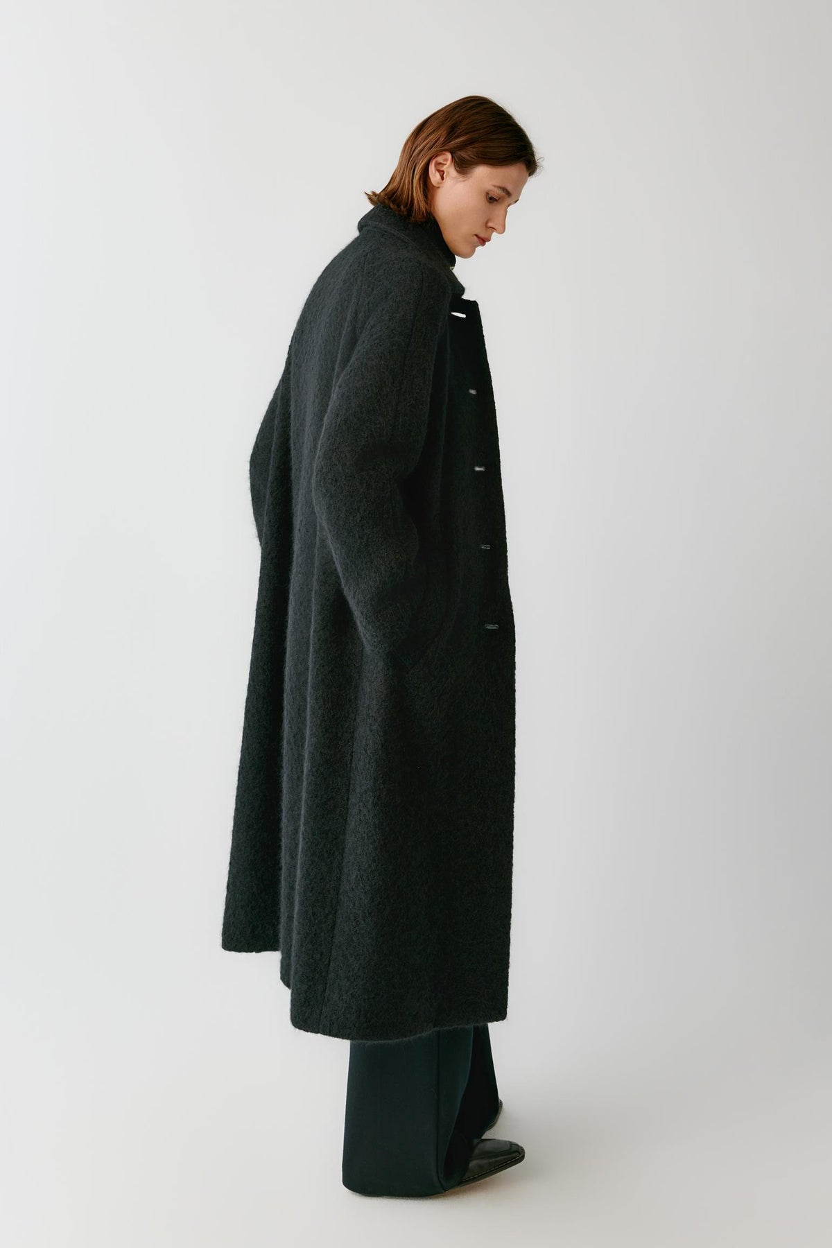 Side view of a woman wearing the deep black alpaca and wool tweed coat, highlighting its long silhouette and textured surface.