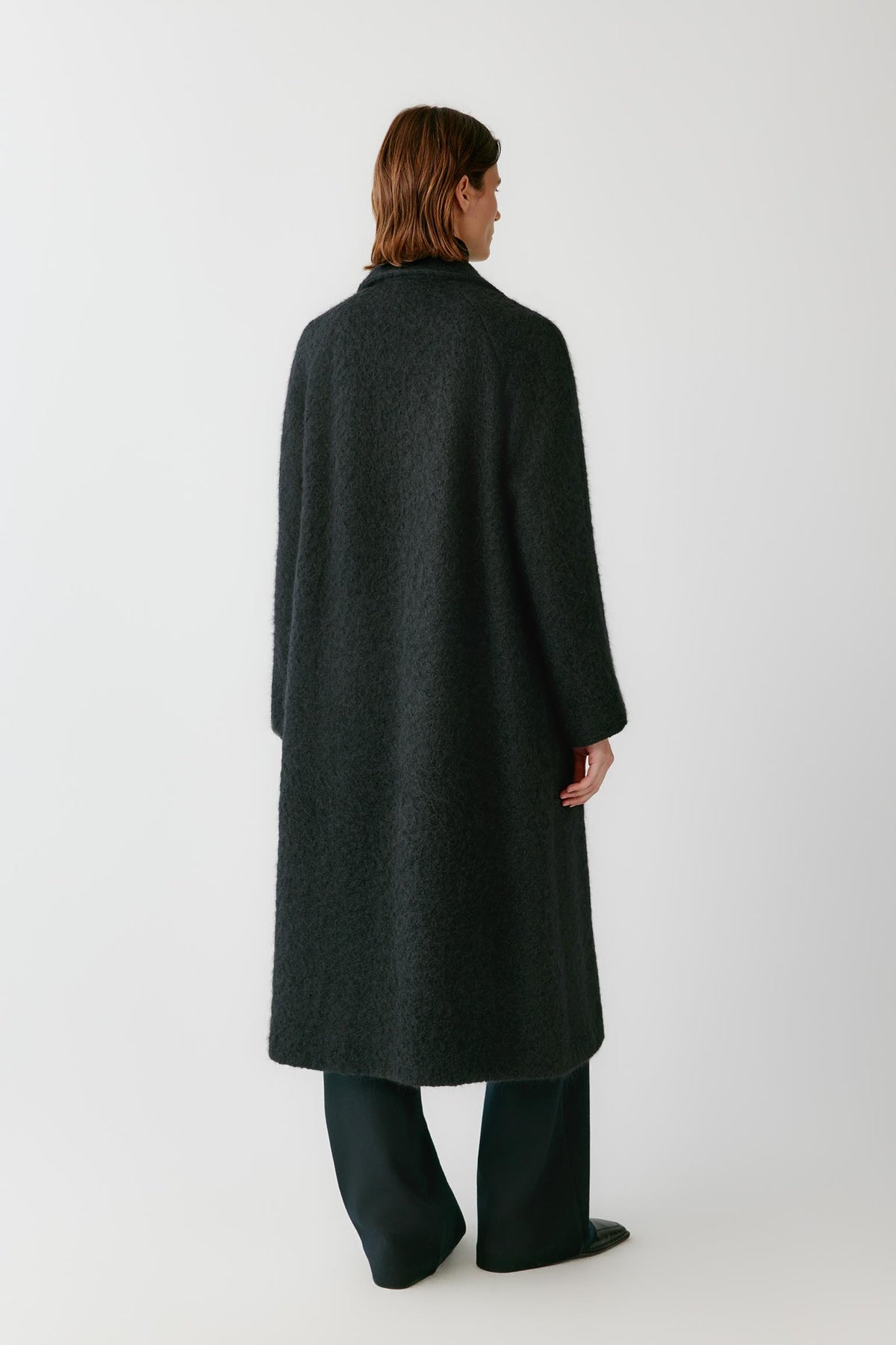 Back view of a woman wearing the deep black alpaca and wool tweed coat, showing the long straight silhouette and dropped shoulders.
