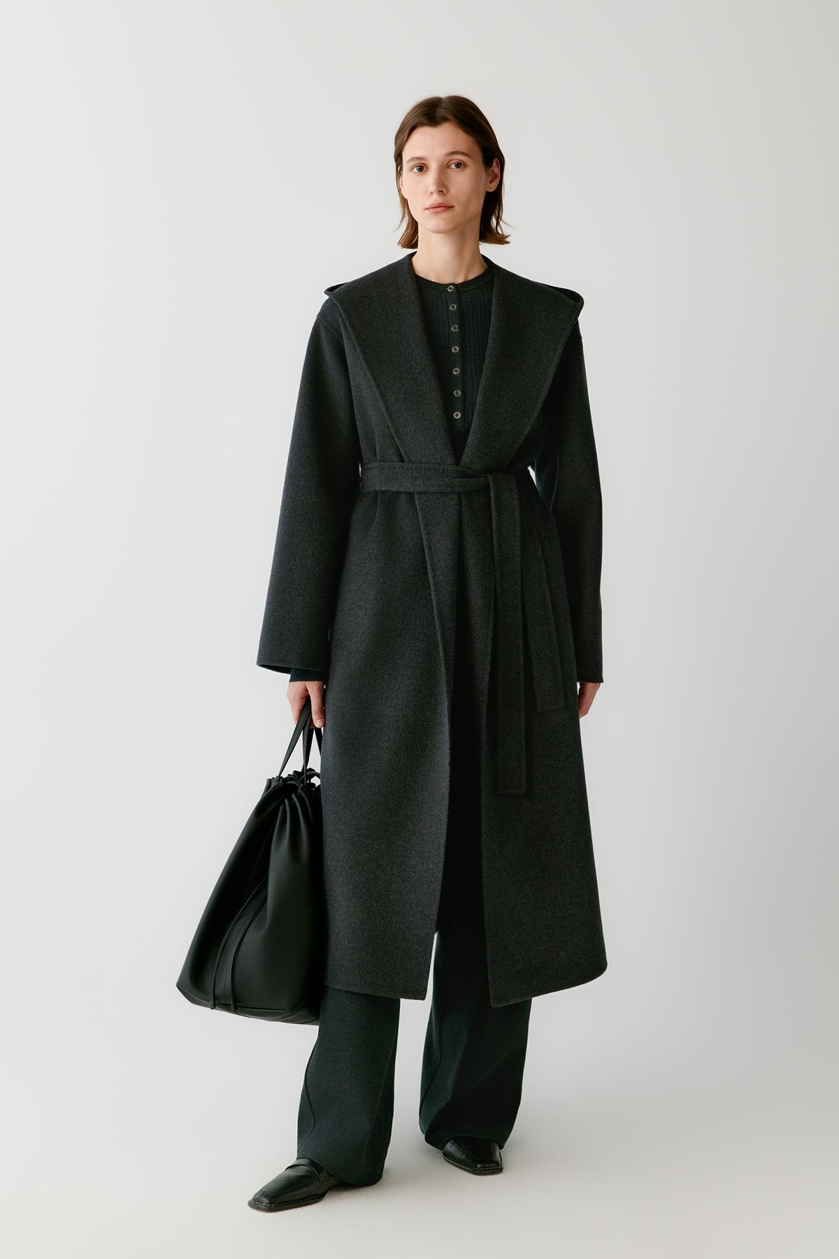 Front view of a woman wearing a dark grey melange double-face wool and cashmere coat, showing the wide shawl collar, self-tie belt and long silhouette.