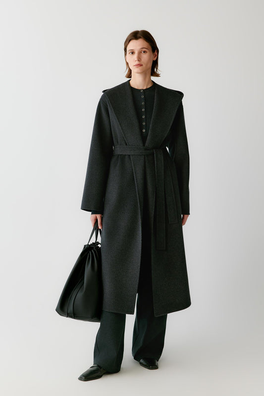 Front view of a woman wearing a dark grey melange double-face wool and cashmere coat, showing the wide shawl collar, self-tie belt and long silhouette.