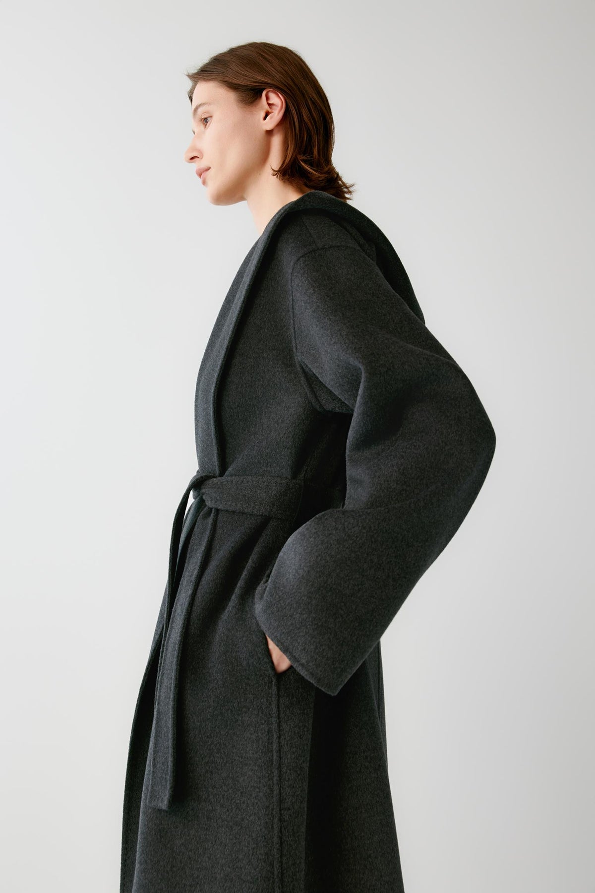 Side view highlighting the long cut, dropped shoulders and structured drape of the dark grey melange coat.