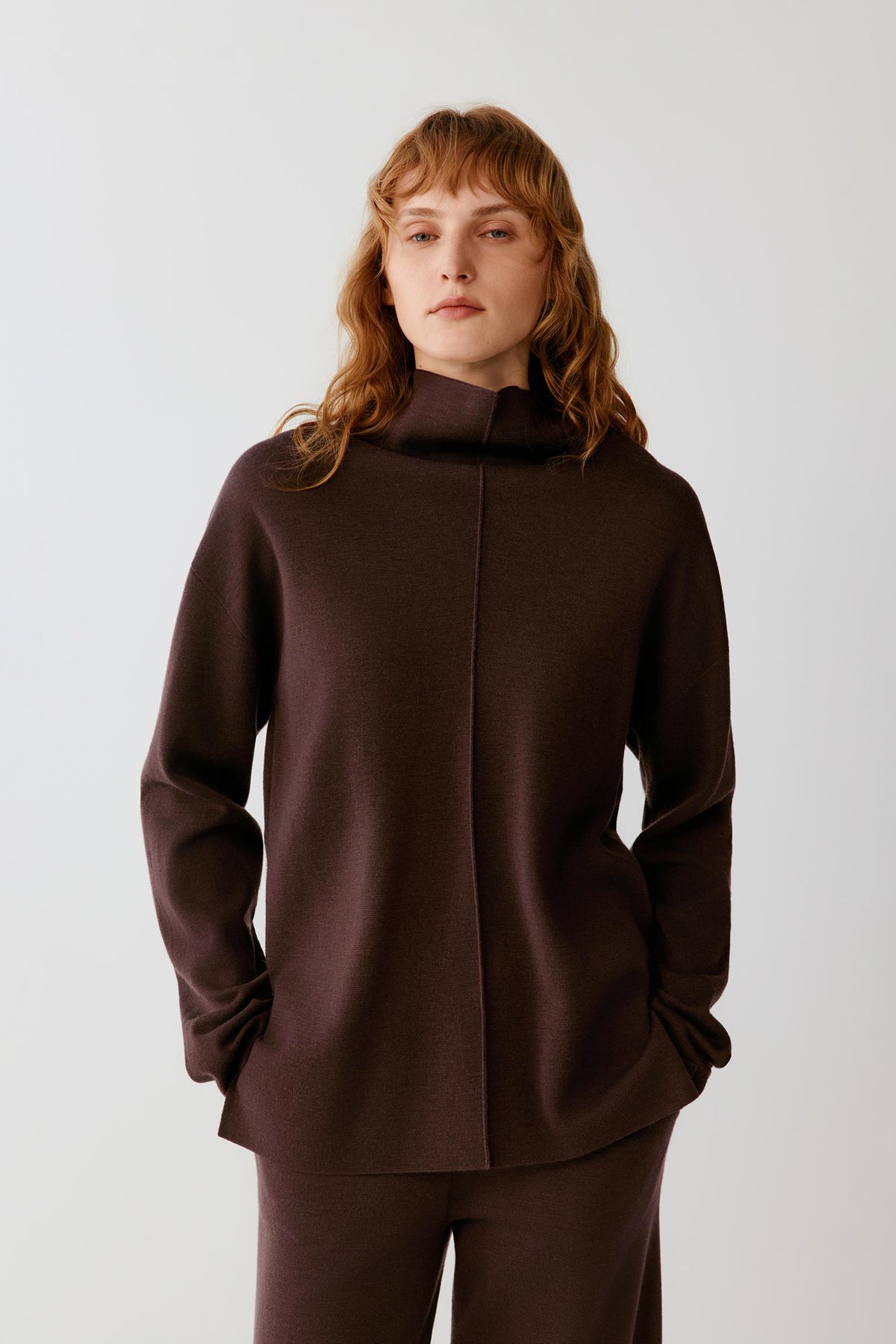Front view of a woman wearing a dark brown merino wool pullover, showing the mock neck, dropped shoulders and straight silhouette.