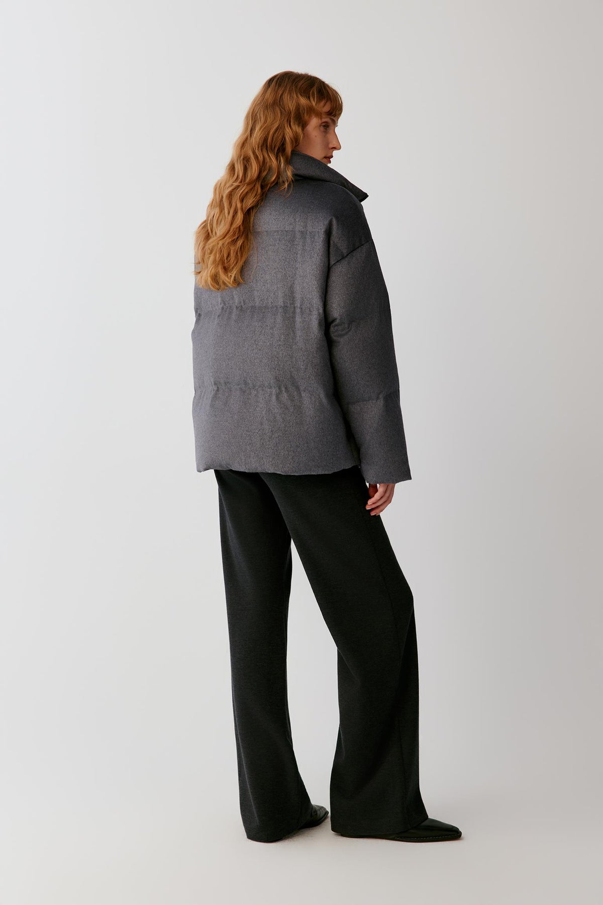 Three-quarter view showing the high stand collar, dropped shoulders and quilted construction of the dark grey melange down coat.