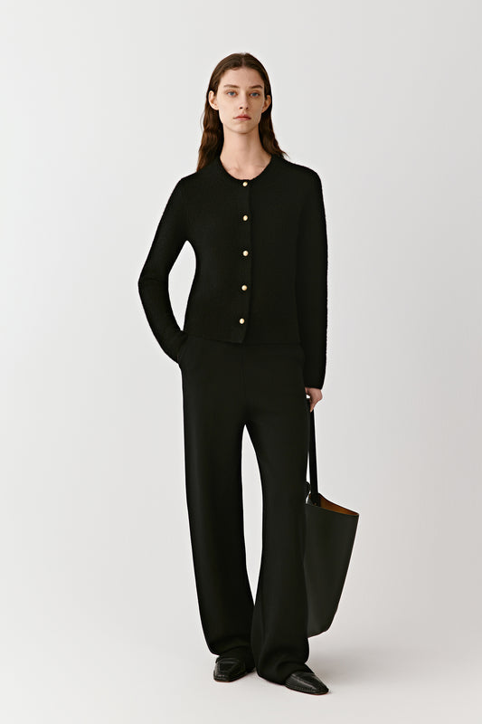 Full-length view of a person wearing a black cashmere cardigan with metal buttons, paired with wide-leg black trousers. The model stands against a light background, hands in pockets, showing the clean, minimal silhouette of the outfit.