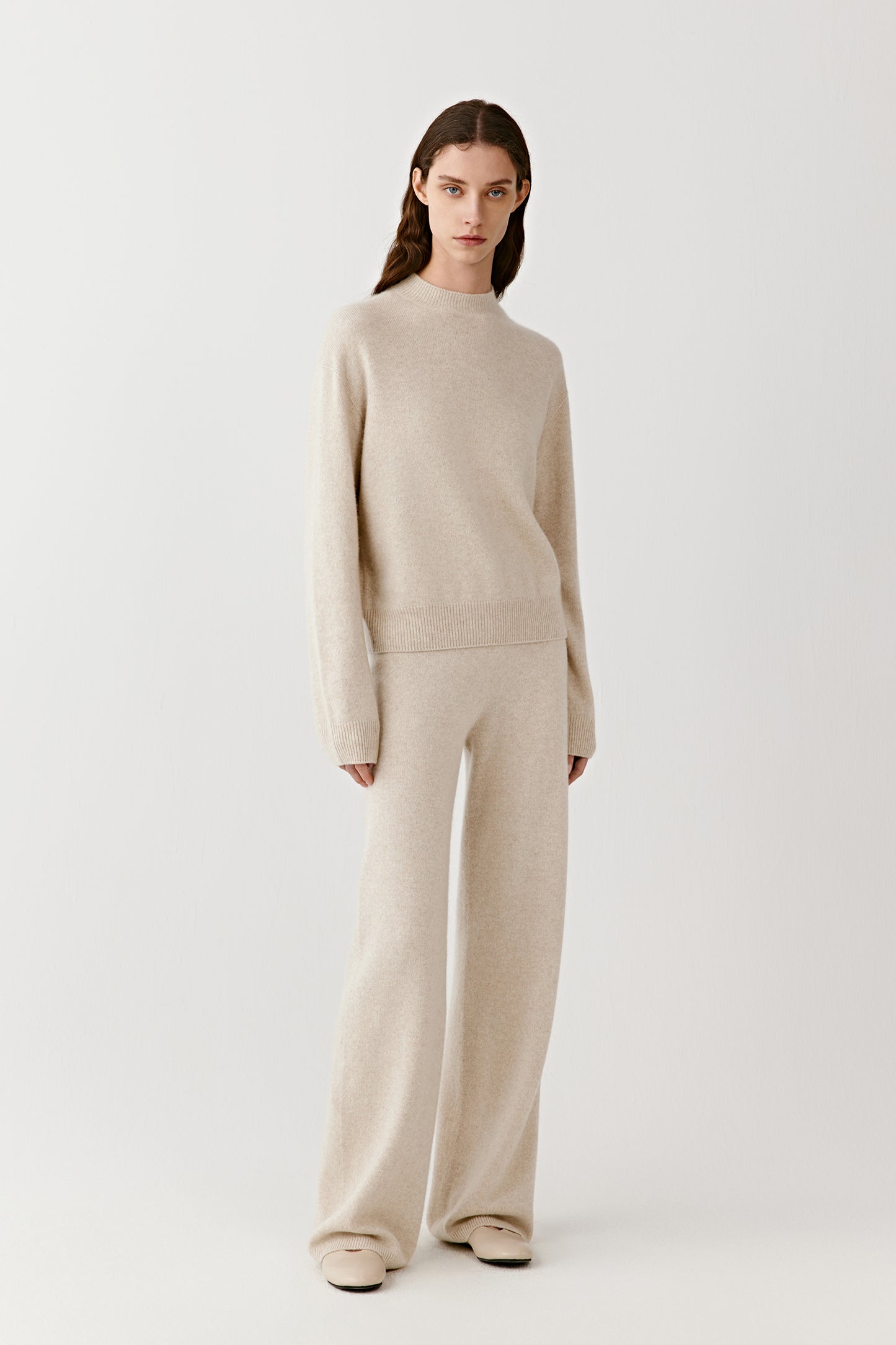 Full-length view of a person wearing cream knitted cashmere trousers with a matching cream sweater, standing against a plain light background.