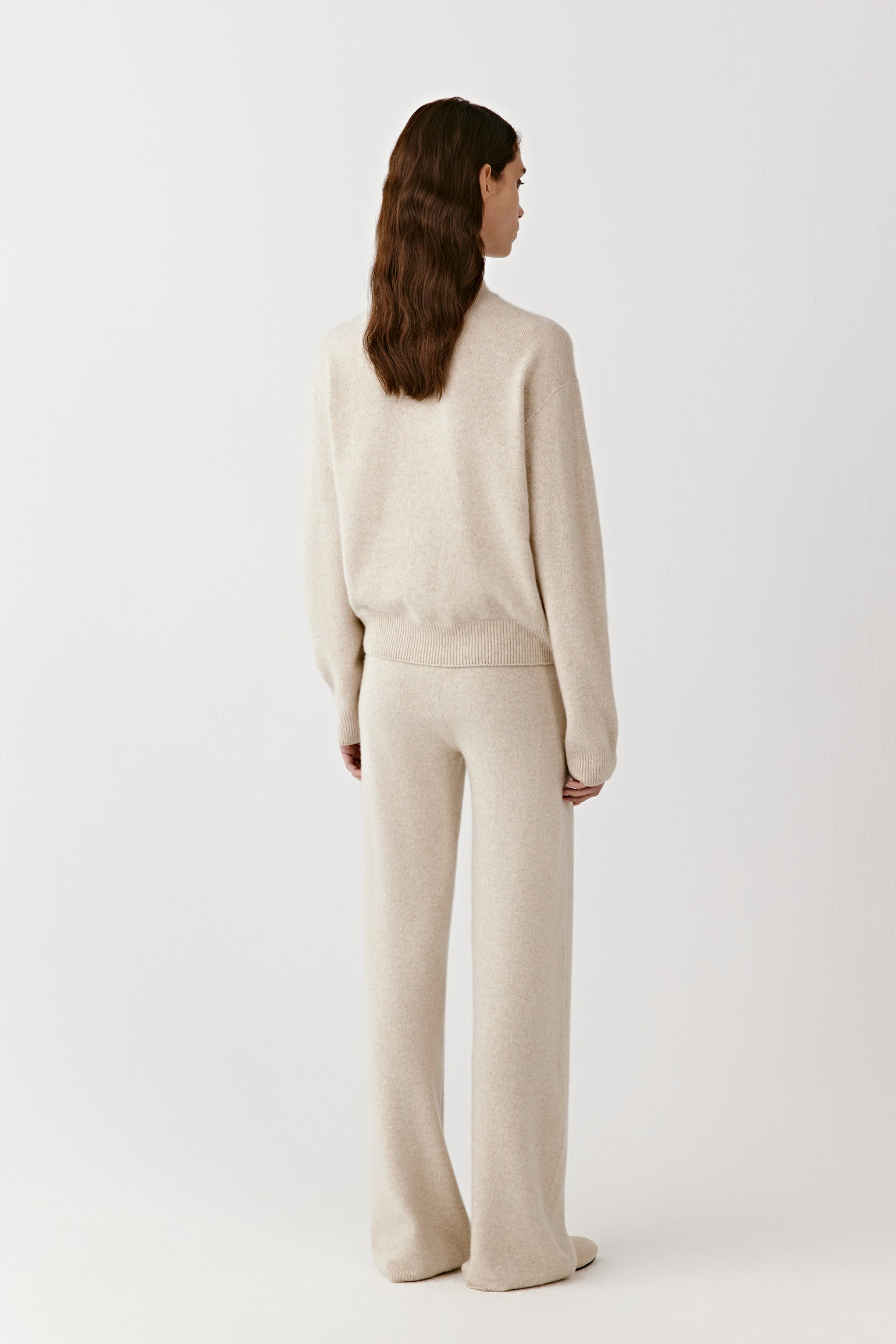Full-body back view of a person wearing cream knitted cashmere trousers and a matching top, highlighting the drape and relaxed fit.