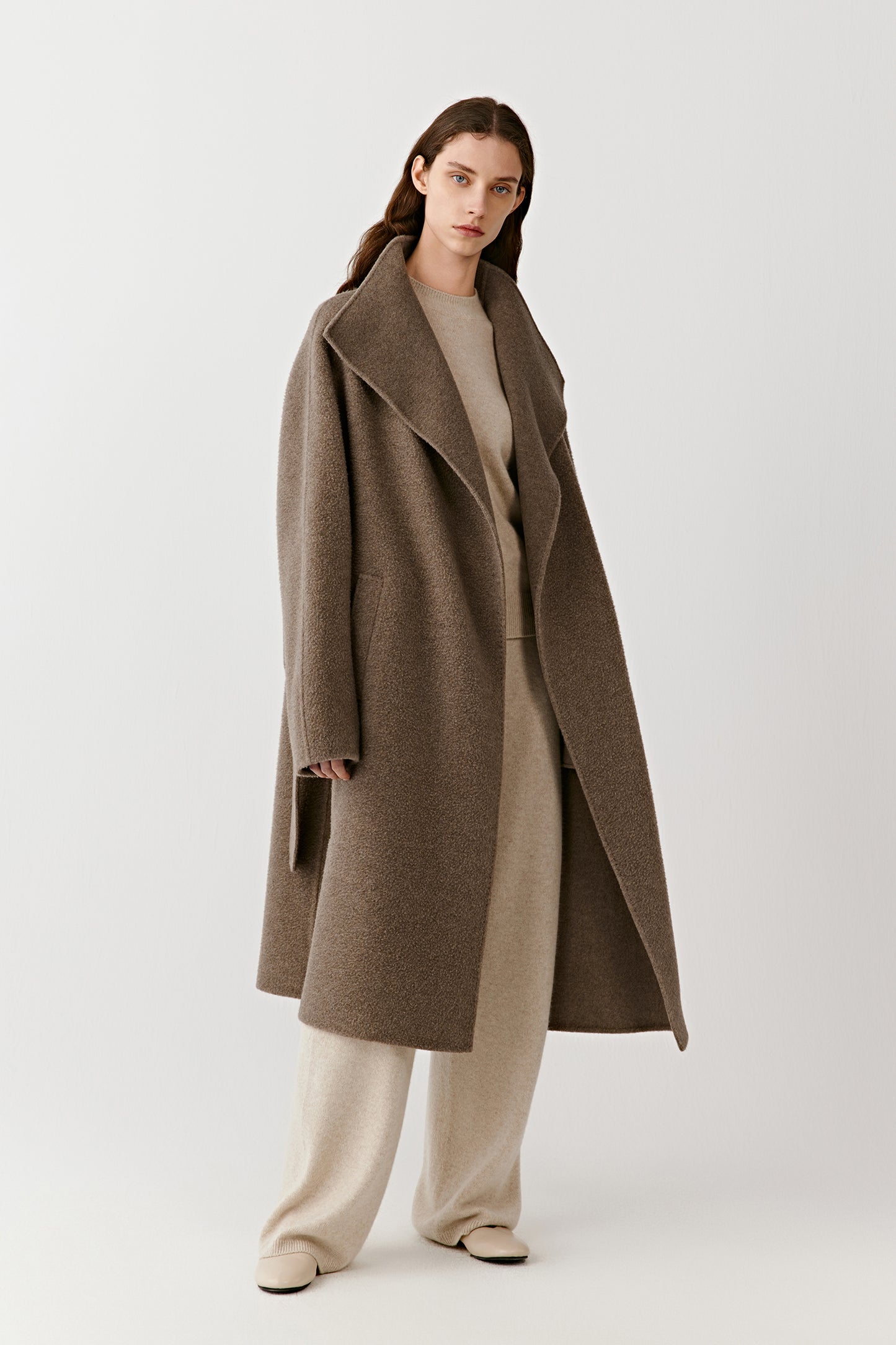 A woman stands facing forward wearing a long wrap coat in undyed double-faced cashmere, styled with light trousers and a soft cream top. The open collar and relaxed belt define the silhouette.