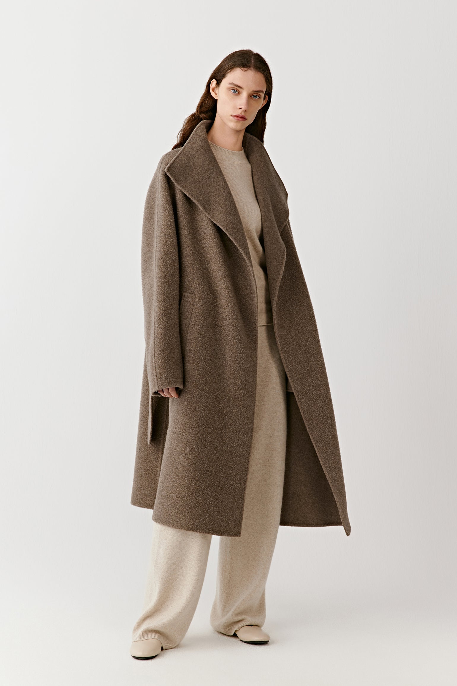 A woman stands facing forward wearing a long wrap coat in undyed double-faced cashmere, styled with light trousers and a soft cream top. The open collar and relaxed belt define the silhouette.