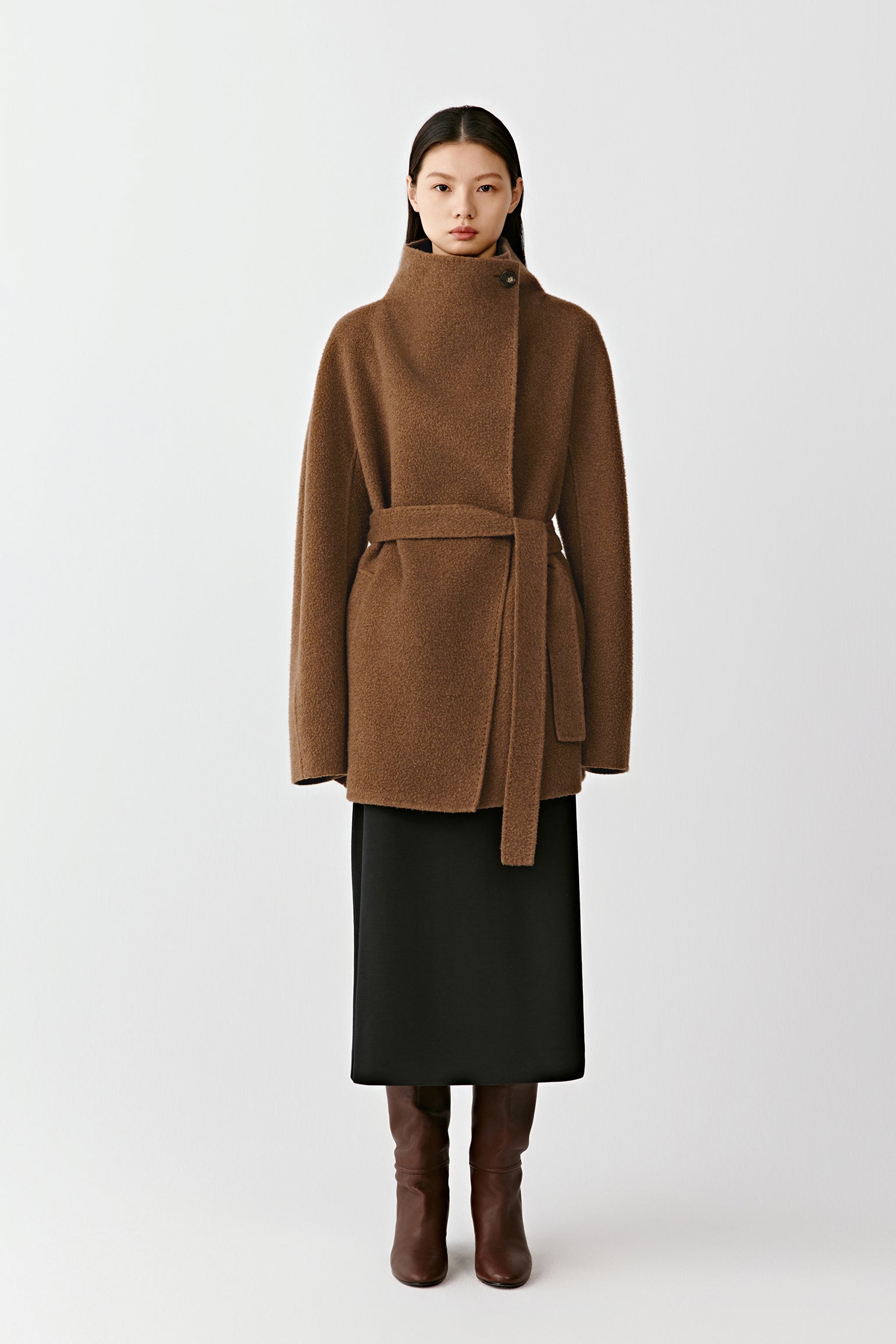 Full-length view of a person wearing a camel and dark brown double-faced cashmere short coat with a wide contrasting collar and a tie belt, paired with a black skirt and tall brown boots. The model stands against a light background.