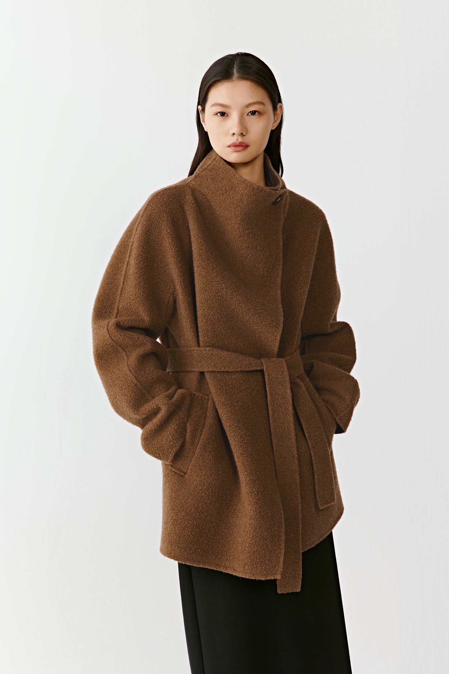 Three-quarter front view of a person wearing the camel and dark brown double-faced cashmere short coat, showing the relaxed shape, patch pockets, and belt tied at the waist. The model stands in front of a neutral backdrop.