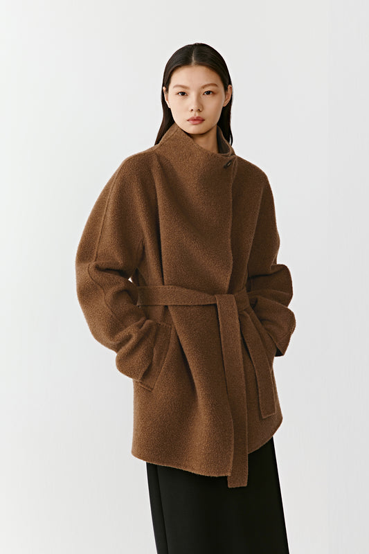 Three-quarter front view of a person wearing the camel and dark brown double-faced cashmere short coat, showing the relaxed shape, patch pockets, and belt tied at the waist. The model stands in front of a neutral backdrop.