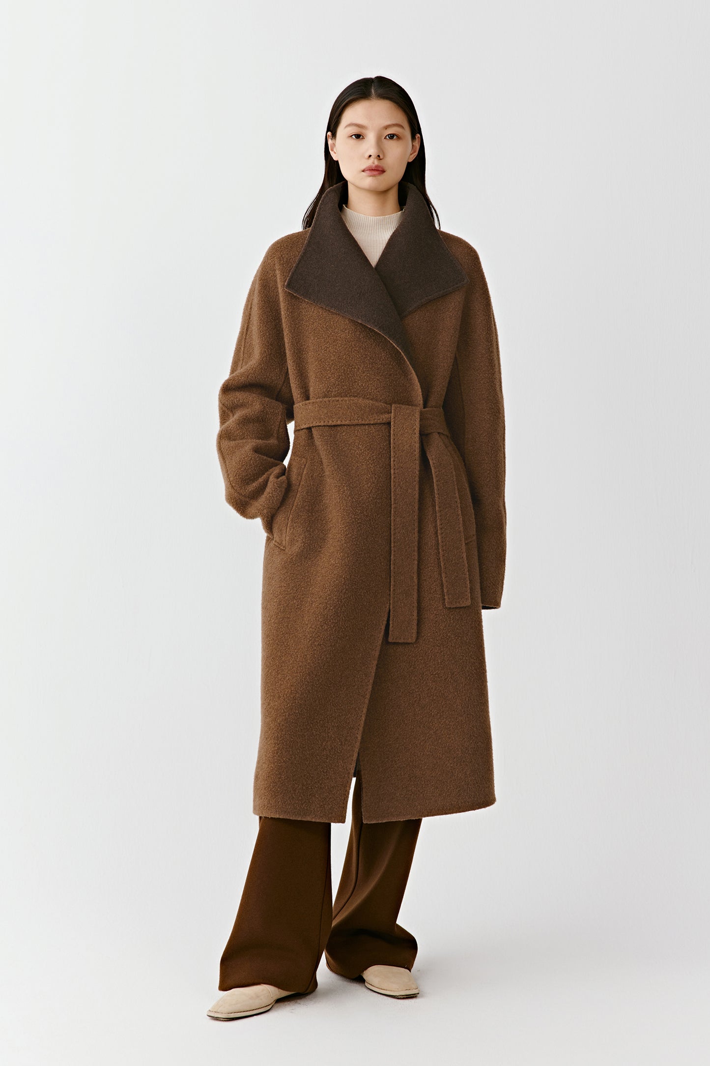 A woman stands facing forward wearing a long two-tone coat in double-faced cashmere, featuring a soft shawl collar and a tied belt, styled with wide-leg trousers.