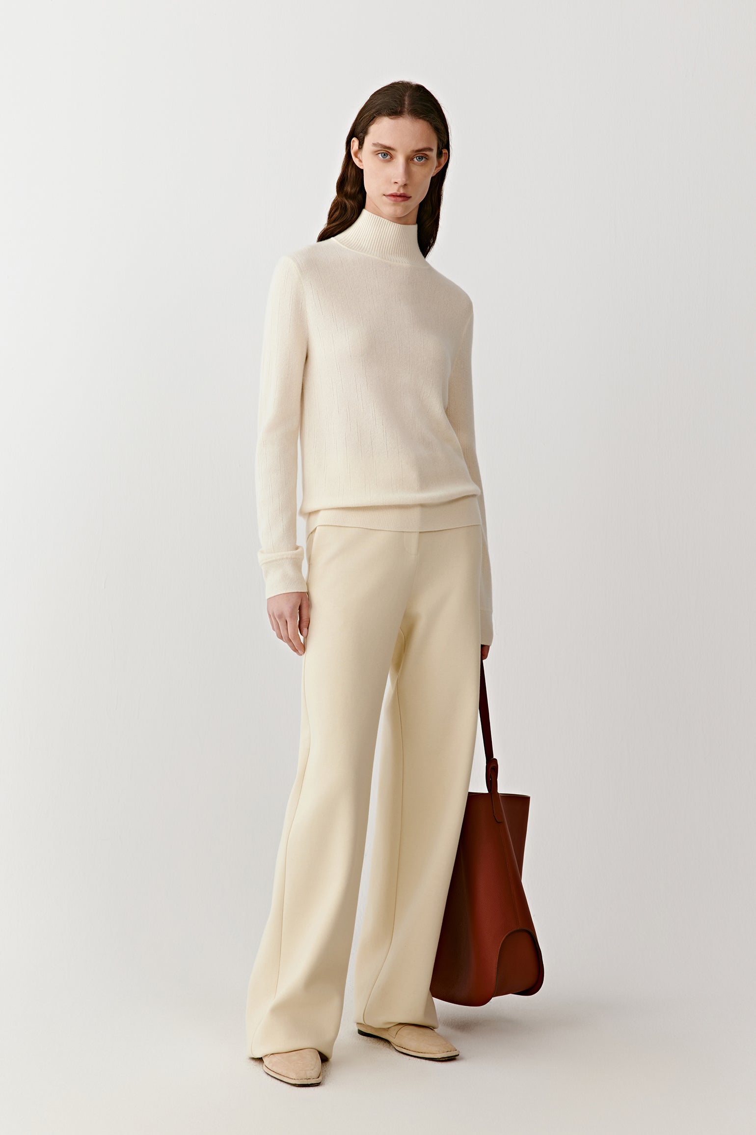 Full-length front view of a cream high-neck cashmere pullover with a fluid drape, paired with matching light trousers.
