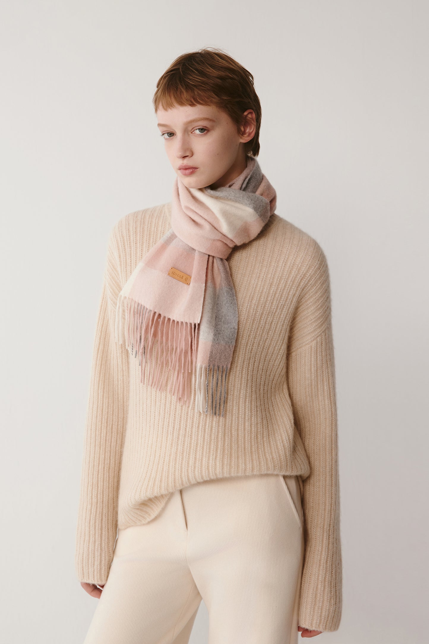 A woman wearing a soft pink checked cashmere scarf draped around her neck, styled over a light beige knit sweater.