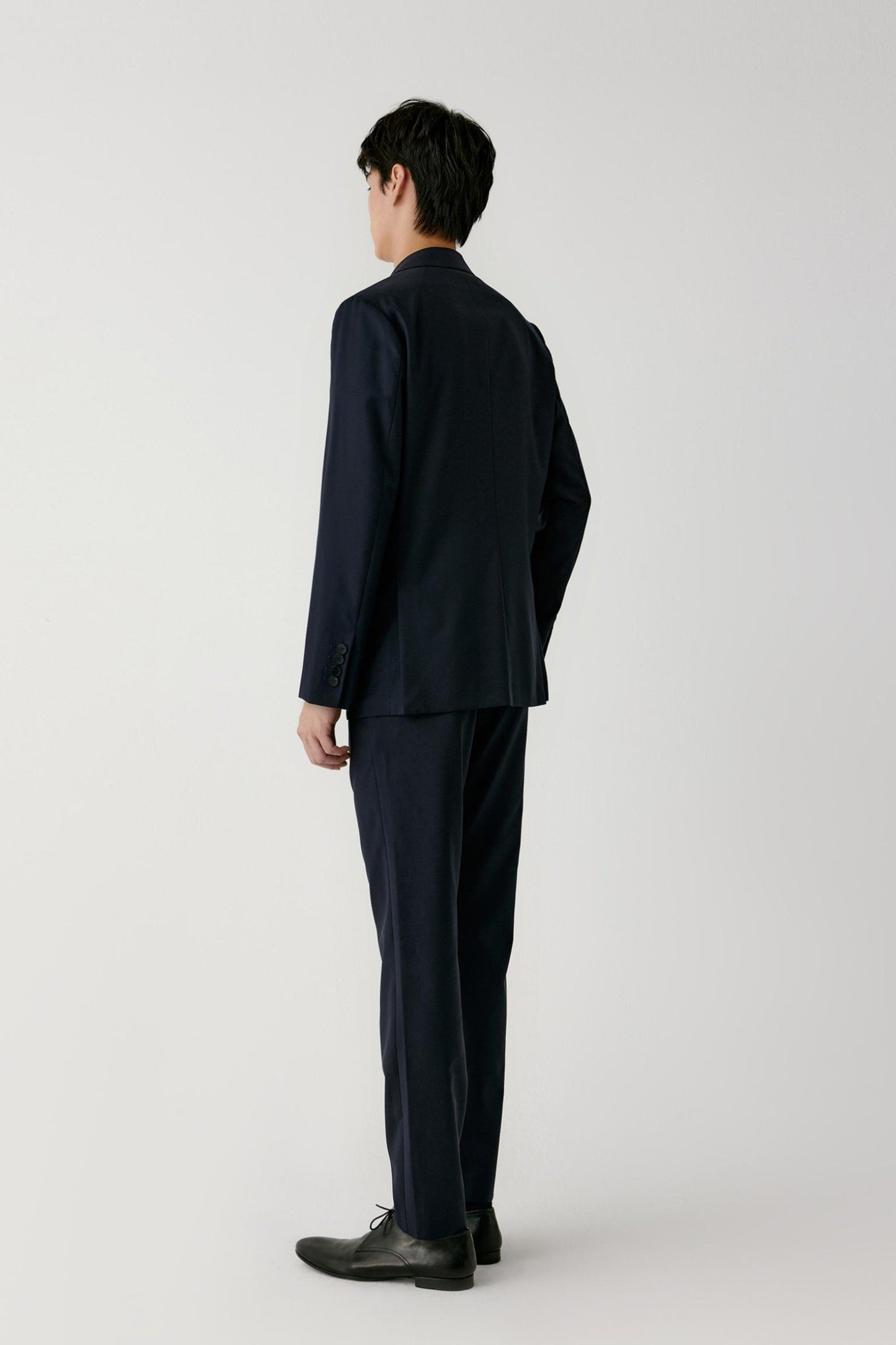 A man wearing a dark navy blue suit, viewed from the back, standing against a plain light gray studio background.