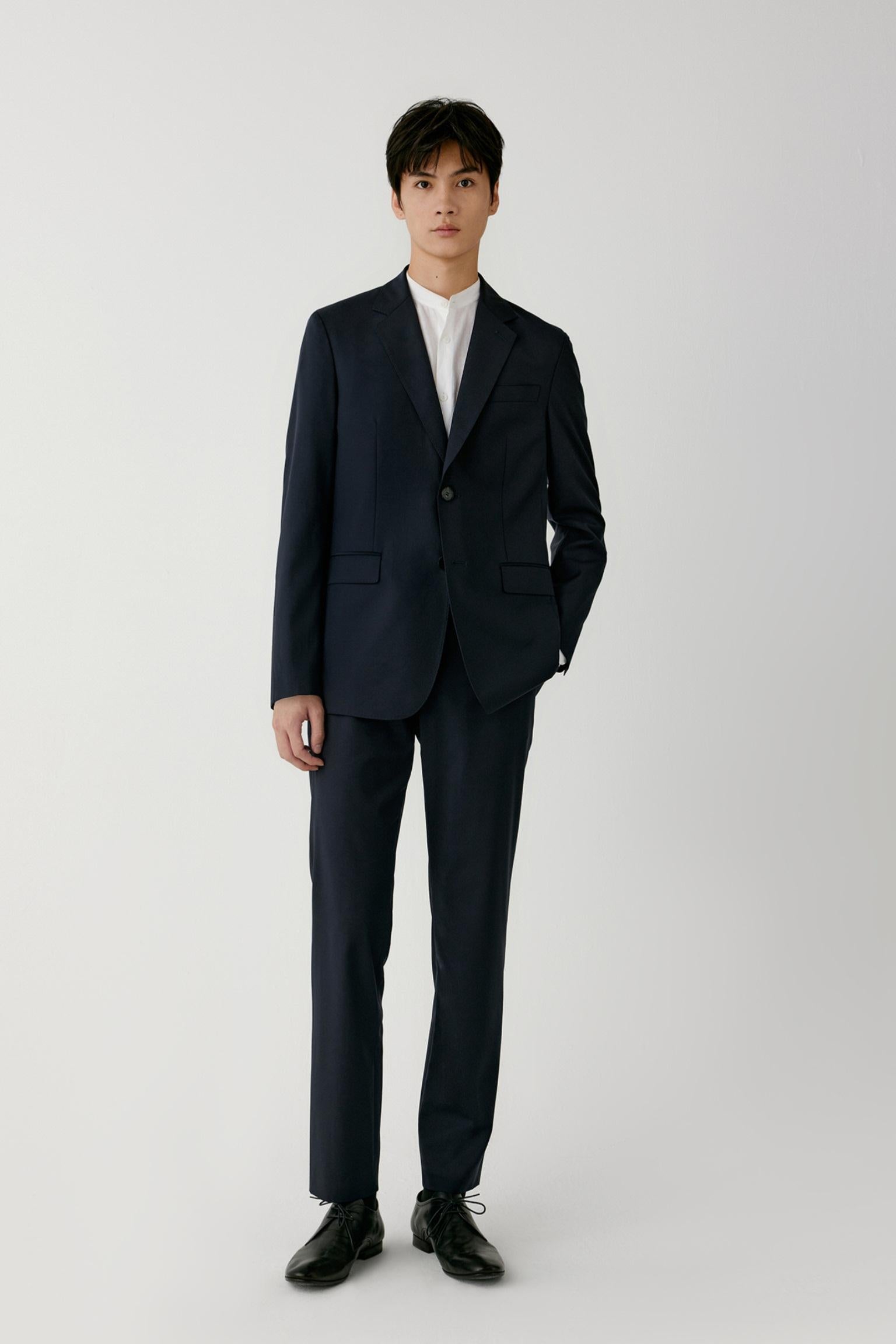 A man modeling a dark navy blue two-piece suit with a white shirt and black dress shoes, standing against a light gray studio background.