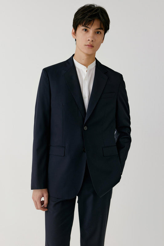 A man wearing a dark navy blue suit jacket and matching trousers over a white collared shirt, standing against a light gray studio background.