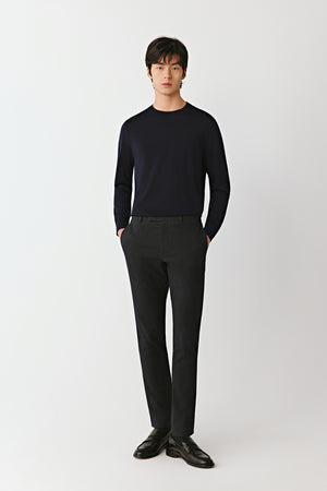 A man wears a blue-black merino wool jumper with dark trousers, standing in front of a pale background for subtle contrast.