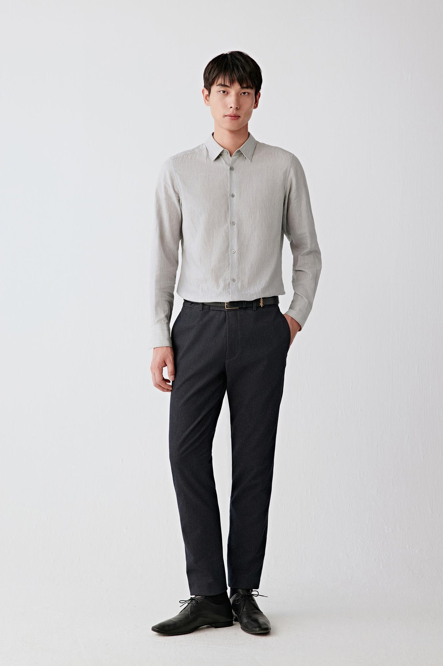 A man modeling a light gray long-sleeve linen button-down shirt paired with dark charcoal trousers and black leather dress shoes, standing against a plain light gray studio background.