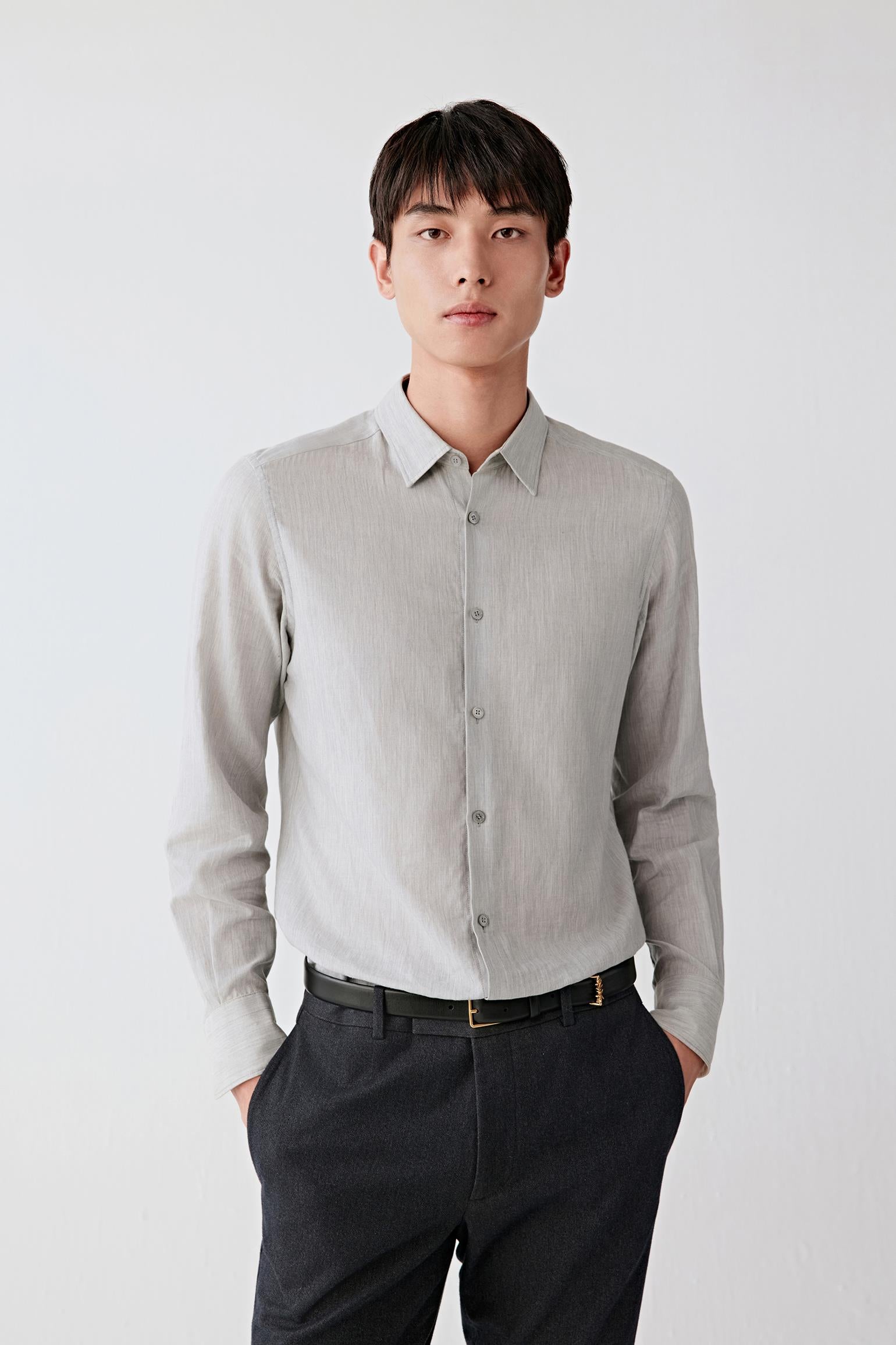 A man wearing a light gray linen button-down shirt and dark charcoal trousers, standing against a plain light gray studio background.