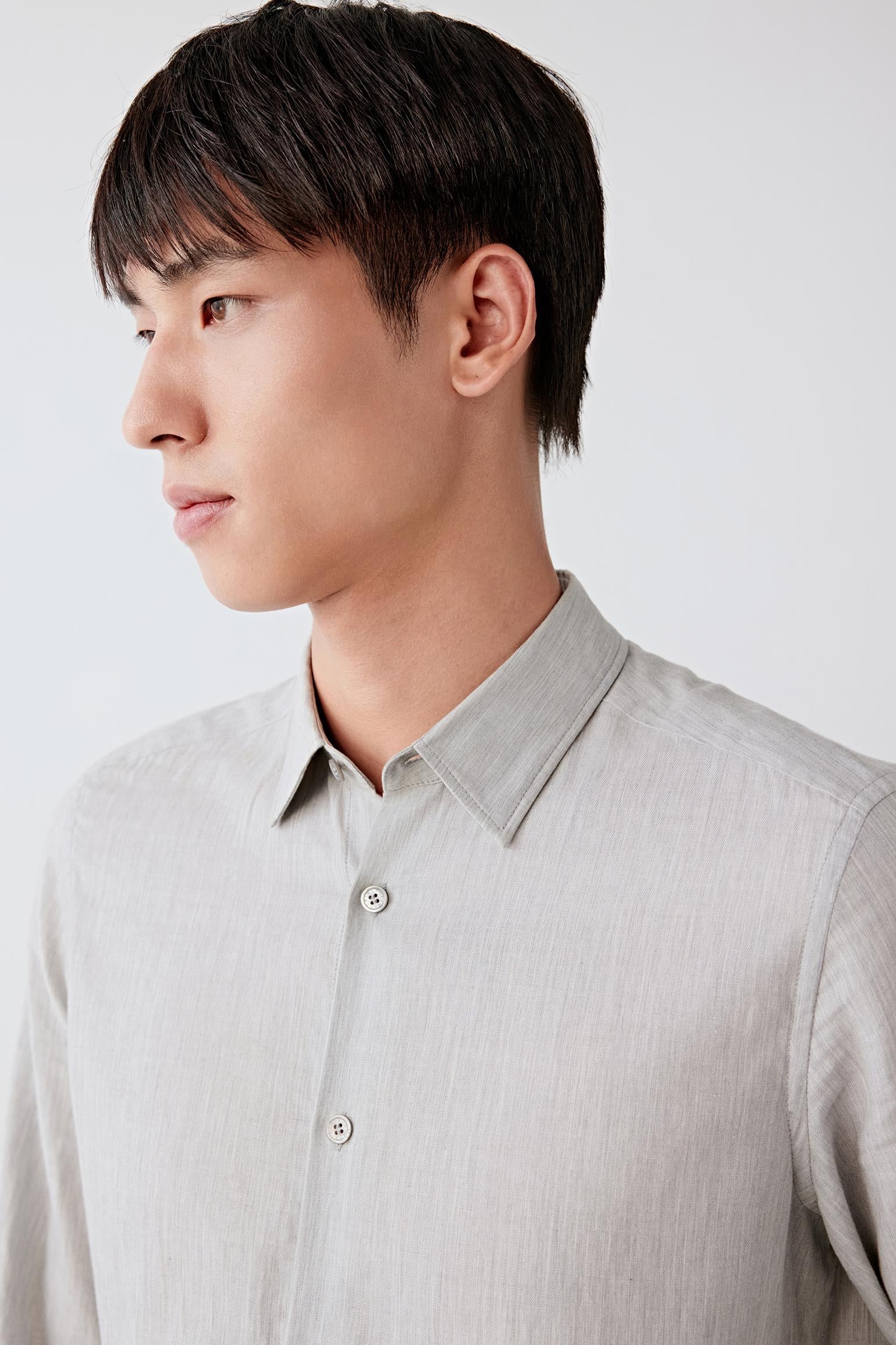 A man in profile wearing a light gray linen button-down shirt, shown from the chest up against a clean, light gray studio background.