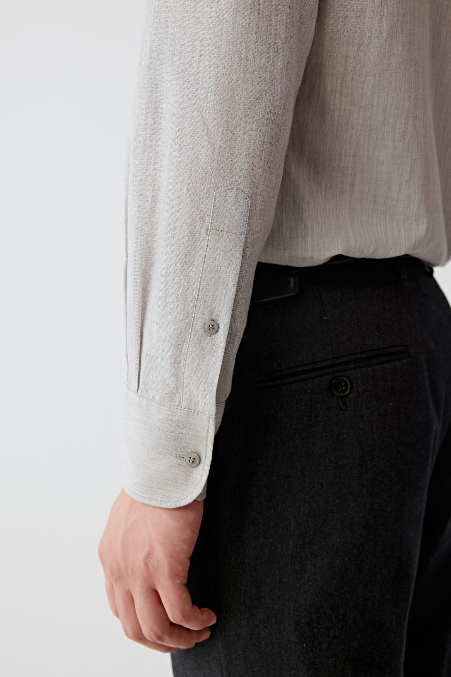Close-up of a person wearing a light beige linen button-up shirt and dark charcoal trousers, showing the sleeve cuff and side pocket detail.