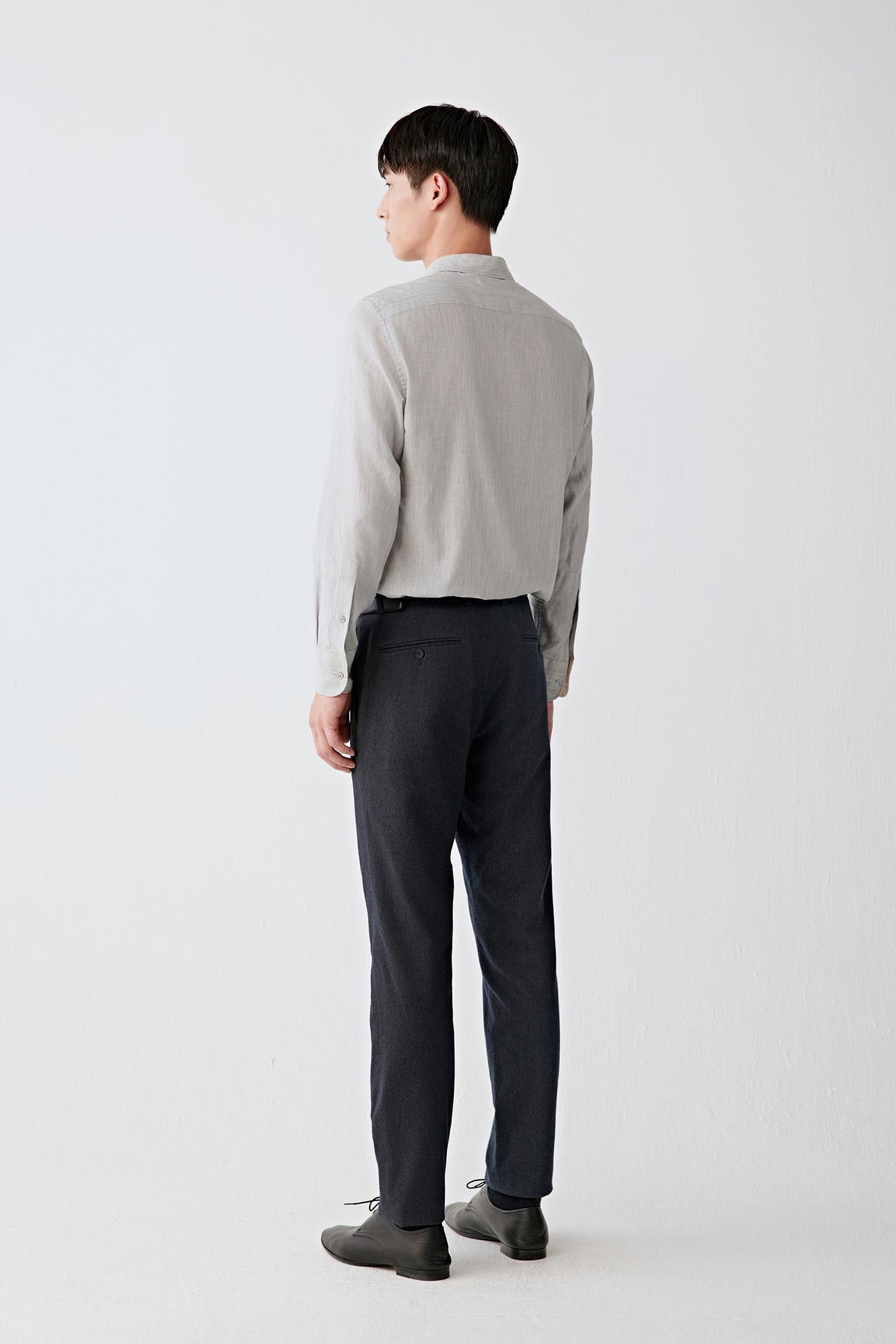 A man shown from the back wearing a light gray long-sleeve shirt and dark charcoal trousers, standing against a plain light gray studio background.