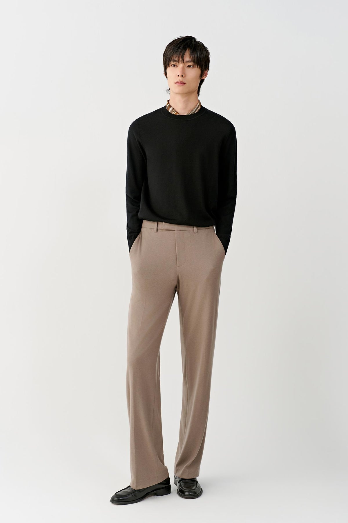 A male model stands against a light gray studio background wearing a black long-sleeve knit top, beige straight-leg trousers, and black leather shoes.